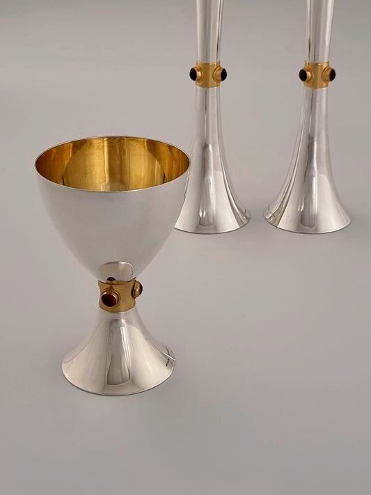 Deborah Kiddush Set. This set was designed in 1993 and is made of silver, gold-plated silver and three garnet stones per stick, and three garnet stones on the cup. The cup is 5” high and the candlesticks are 7½“ high.